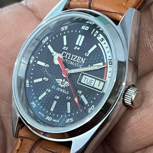 May include: A Citizen automatic wristwatch with a dark blue face, silver case, and brown leather strap. The watch face displays the day and date, with white and red hands. The watch is marked "21 Jewels" and "Japan 30 Made".