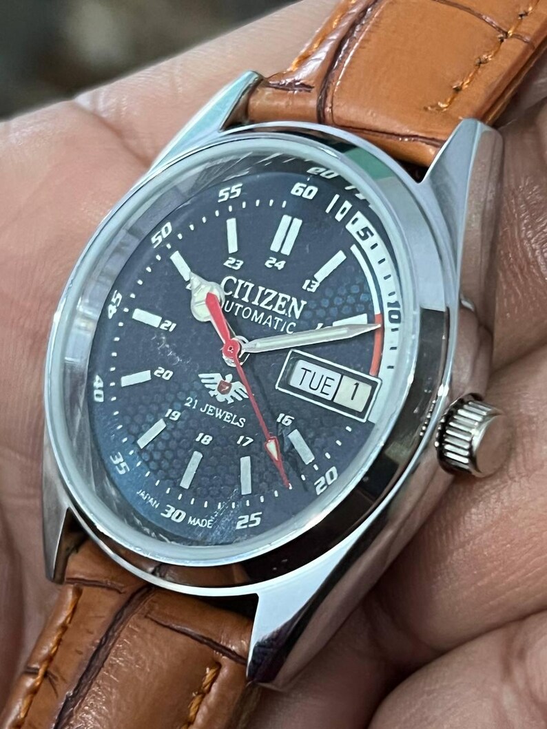 May include: A Citizen automatic wristwatch with a dark blue face, silver case, and brown leather strap. The watch face displays the day and date, with white and red hands. The watch is marked "21 Jewels" and "Japan 30 Made".