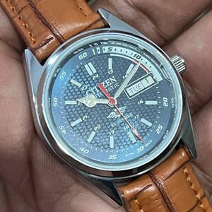 May include: A Citizen automatic wristwatch with a dark blue dial and a brown leather strap. The watch features a day and date display and a red second hand. The watch face has a textured pattern and silver-tone hour markers.