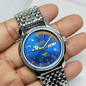 May include: A Citizen automatic wristwatch with a blue dial and silver-tone stainless steel band. The watch face displays the day of the week and has gold-tone hands and markers. The watch is marked "21 Jewels" and "Japan Made".