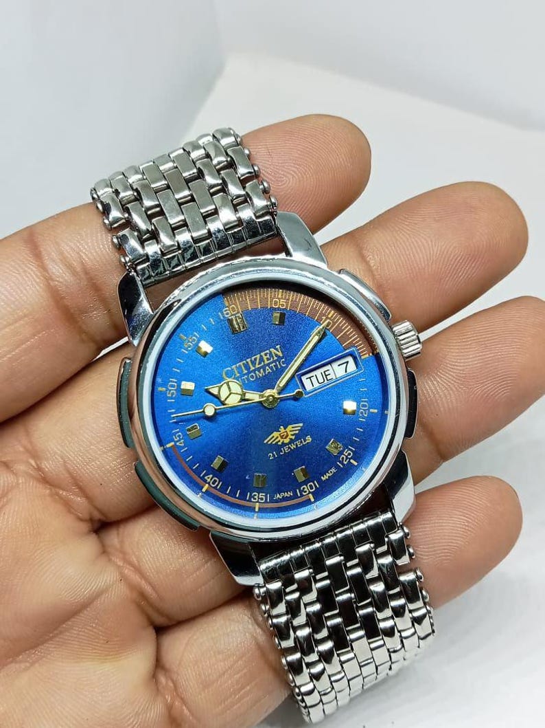 May include: A Citizen automatic wristwatch with a blue dial and silver-tone stainless steel band. The watch face displays the day of the week and has gold-tone hands and markers. The watch is marked "21 Jewels" and "Japan Made".