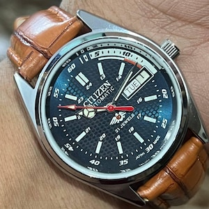 May include: A Citizen automatic watch with a dark blue face and a brown leather strap. The watch face displays the day and date, with white and red accents. The watch has a silver-coloured metal casing.