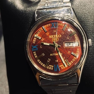 Puede incluir: Un reloj de pulsera automático Seiko vintage con una correa plateada y una esfera marrón rojiza. La esfera del reloj presenta marcadores de hora dorados, números romanos azules y la visualización del día y la fecha. El reloj se muestra sobre una superficie negra.