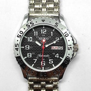 Op de afbeelding: Zilverkleurig Seiko automatisch horloge met een zwarte textuur wijzerplaat. Het horloge heeft witte cijfers, een rode secondewijzer en een dag/datum weergave. De horlogeband is een zilveren schakelarmband.