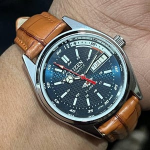 May include: A Citizen automatic wristwatch with a dark blue face, silver case, and brown leather strap. The watch face displays the day of the week and the time. The watch has red hands and a textured dial.