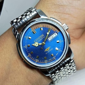 May include: A Citizen automatic wristwatch with a blue dial and silver-tone metal band. The watch face displays the day of the week and the time. The watch has a silver-tone case and a textured metal band.