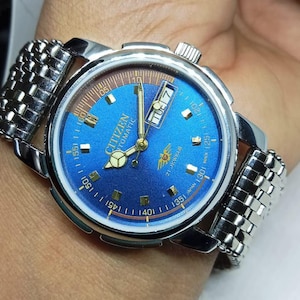 May include: A Citizen automatic wristwatch with a blue face and silver-tone metal band. The watch face displays the day and date, with gold-tone hands and markers. The watch is on a wrist.