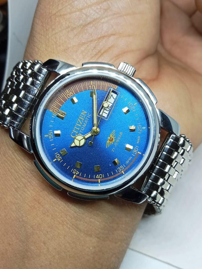 May include: A Citizen automatic wristwatch with a blue face and silver-tone metal band. The watch face displays the day and date, with gold-tone hands and markers. The watch is on a wrist.