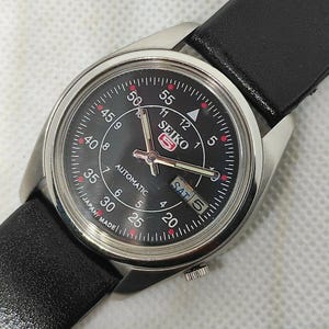 Könnte beinhalten: Nahaufnahme einer automatischen Seiko-Armbanduhr mit schwarzem Zifferblatt und silberfarbenem Gehäuse. Die Uhr hat ein schwarzes Lederarmband und zeigt den Wochentag an. Das Zifferblatt hat weiße und rote Markierungen.