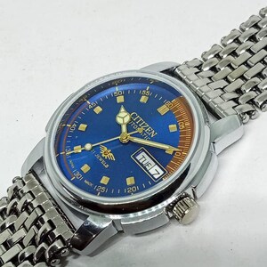 May include: A vintage Citizen automatic wristwatch with a blue dial and silver-tone metal band. The watch face displays the day of the week and the text "Citizen Automatic 21 Jewels". The watch has a silver-tone case and a textured metal band.