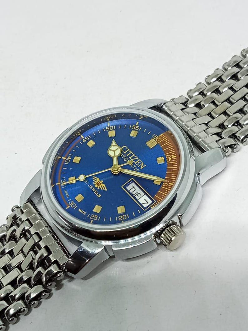 May include: A vintage Citizen automatic wristwatch with a blue dial and silver-tone metal band. The watch face displays the day of the week and the text "Citizen Automatic 21 Jewels". The watch has a silver-tone case and a textured metal band.