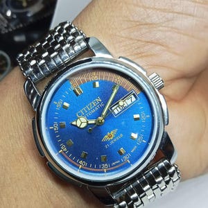 May include: A Citizen automatic wristwatch with a blue dial and a silver-tone stainless steel band. The watch face features gold-tone hands and markers, a day-date display, and the text "Citizen Automatic 21 Jewels Japan Made".