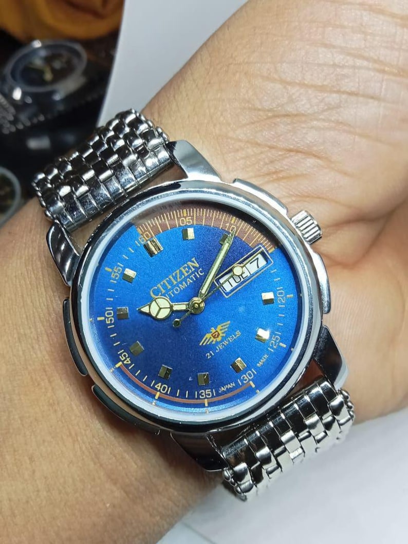 May include: A Citizen automatic wristwatch with a blue dial and a silver-tone stainless steel band. The watch face features gold-tone hands and markers, a day-date display, and the text "Citizen Automatic 21 Jewels Japan Made".