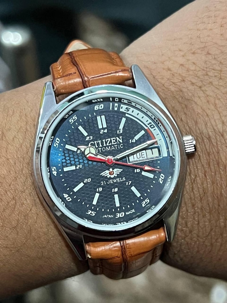 May include: A Citizen automatic wristwatch with a dark blue face, silver bezel, and brown leather strap. The watch face displays the day and date, with a red second hand. The watch is marked "21 Jewels" and "Japan 30 Made".