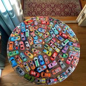 Könnte beinhalten: Ein runder Tisch mit einer transparenten Harzplatte, in die zahlreiche Spielzeug-Rennwagen in verschiedenen Farben eingebettet sind. Die Autos sind dicht angeordnet und bilden ein lebendiges und verspieltes Design. Der Tisch steht auf einem Holzboden.