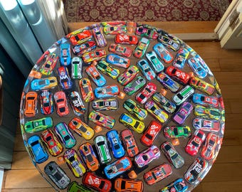 May include: A round table with a clear resin top embedded with numerous toy race cars in various colors. The cars are arranged densely, creating a vibrant and playful design. The table sits on a wooden floor.