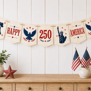 May include: A patriotic banner with the words "HAPPY AMERICA 250" and images of the Liberty Bell, an eagle, and the Statue of Liberty. The banner is strung with twine and displayed above a wooden surface with American flags and a star.