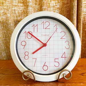 Peut inclure: Une horloge à quartz Cardinal blanche avec une lunette noire et des chiffres rouges. Le cadran de l'horloge a un motif quadrillé et les aiguilles sont rouges. L'horloge est présentée sur un support en métal doré.