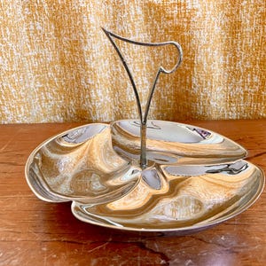 May include: A silver-coloured, three-section serving dish with a handle. The dish has an organic shape with a reflective surface, suitable for serving snacks. The handle is a decorative element. The dish is made of metal.