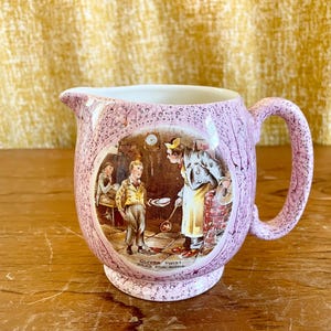May include: A vintage pink ceramic jug with a handle and spout. The jug has a detailed illustration of a scene from Oliver Twist. The text "Oliver Twist asks for more" is visible on the base of the jug.