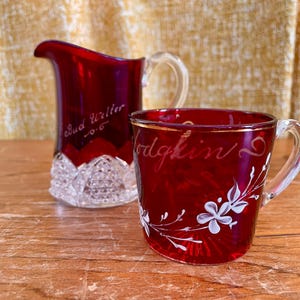 May include: A red glass jug and mug set. The jug has a clear handle and a cut-glass base. The mug has a clear handle and white floral accents. Both pieces have handwritten text. The jug reads "Bud Writer" and the mug reads "Hodgkin".
