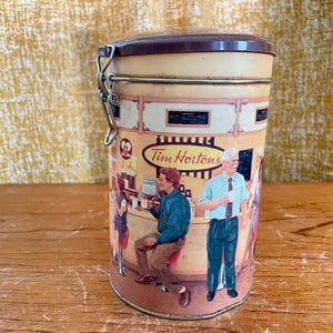 May include: A cylindrical metal container with a lid and clasp, featuring a vintage Tim Hortons design. The illustration depicts people in a coffee shop setting, with the Tim Hortons logo prominently displayed.