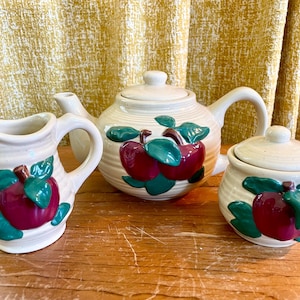 May include: A cream ceramic tea set comprising a teapot, creamer jug, and sugar bowl. Each item features a red apple design with green leaves. The set is displayed on a wooden surface.