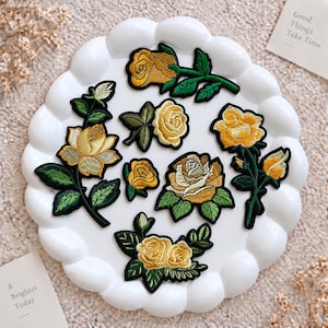 Puede incluir: Una colección de siete parches bordados de rosas amarillas con hojas y tallos verdes, dispuestos sobre un plato blanco festoneado. Los parches presentan varios tamaños y orientaciones de rosas, detallados con hilos intrincados y bordes negros.