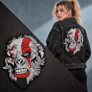 Könnte beinhalten: Eine schwarze Jeansjacke mit einem großen gestickten Aufnäher auf der Rückseite. Der Aufnäher zeigt ein stilisiertes weißes Gorillagesicht mit leuchtend blauen Augen und einem markanten roten Streifen, der vertikal über ein Auge verläuft.