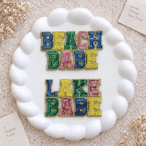 Può includere: Due toppe rettangolari termoadesive con paillettes multicolori. La toppa superiore mostra il testo BEACH BABE in lettere blu, gialle, verdi e rosa. La toppa inferiore mostra il testo LAKE BABE in lettere blu, gialle, verdi e rosa.