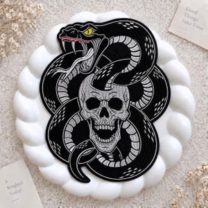 May include: An embroidered patch featuring a black and silver snake coiled around a human skull. The snake has yellow eyes and a pink tongue, while the skull is depicted with an open mouth. The design is rendered in detailed thread work.