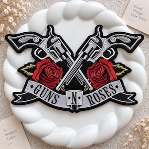 May include: An embroidered patch featuring two crossed silver revolvers and two red roses with green leaves. A light gray banner across the bottom displays the black text GUNS N ROSES. The design is set against a black background with a thick border.