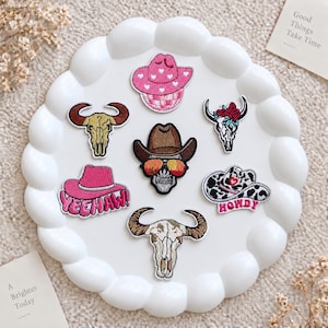 May include: A collection of seven embroidered patches featuring western themes, including a pink cowboy hat, a skull with a brown hat and sunglasses, a cow skull, a pink hat with the text YEEHAW, and a cow print hat with the text HOWDY.
