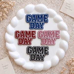 Może przedstawiać: Cztery błyszczące naszywki termoprzylepne z napisem GAME DAY czcionką w stylu uniwersyteckim. Naszywki są dostępne w kolorze niebieskim, ciemnoczerwonym, jasnoróżowym i czarnym, każda z białą obwódką, ułożone na białym talerzu.