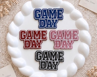 Puede incluir: Cuatro parches termoadhesivos con purpurina que muestran el texto GAME DAY en una fuente de estilo universitario. Los parches están disponibles en azul, rojo oscuro, rosa claro y negro, cada uno con un borde blanco, dispuestos en un plato.