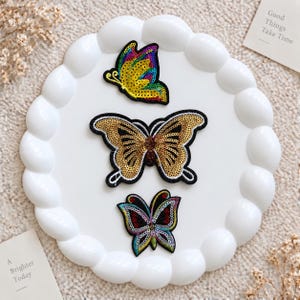 May include: Three butterfly-shaped embroidered patches with sequin detailing. The top patch is yellow, blue, and purple. The centre patch is gold with black outlines. The bottom patch features red, blue, and green tones with black accents.