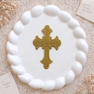 Puede incluir: Un parche en forma de cruz de color dorado con un diseño intrincado y ornamentado y un acabado texturizado brillante. La cruz presenta patrones detallados de estilo filigrana en toda su superficie, centrada en un plato circular blanco.