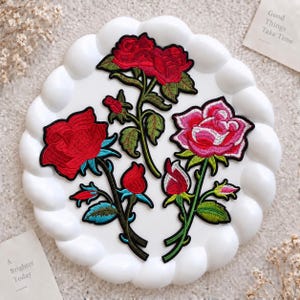 May include: Three embroidered rose patches featuring detailed red and pink flower heads with green stems and leaves. The designs include two fully bloomed roses, one pink rose, and several small rosebuds, all outlined in black thread.