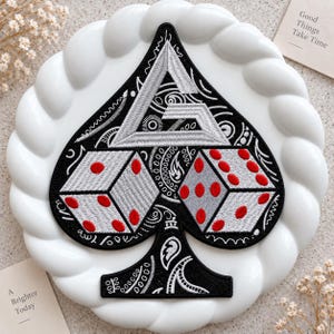 May include: A black and white embroidered patch in the shape of a spade playing card suit. The design features a large letter G at the top, two dice with red dots in the center, and intricate white paisley patterns on a black background.