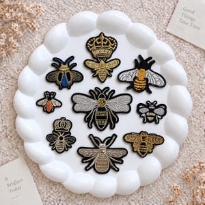 May include: A collection of nine embroidered bee patches in various designs, colors, and sizes, including gold, silver, blue, and black threads, arranged on a white scalloped plate. Two small cards with the text Good Things Take Time and A Brighter Today are visible.