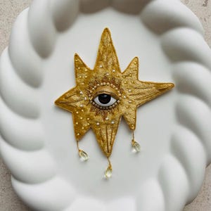 May include: Gold star-shaped brooch featuring an eye design. The brooch is adorned with small white beads and clear, teardrop-shaped crystals. The eye has a black pupil and a brown iris. The brooch is displayed on a white, textured surface.