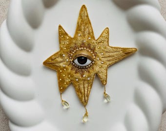 Op de afbeelding: Een goudkleurige, stervormige broche met een oogontwerp in het midden. De broche is versierd met kleine witte kralen en bungelende, druppelvormige, heldere kristallen. Het oog heeft een zwarte pupil en een bruine iris.