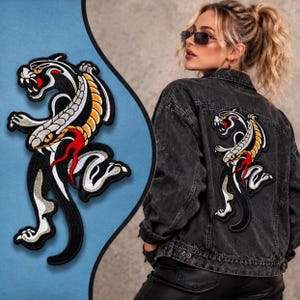 Può includere: Una giacca di jeans nera con una toppa ricamata sul retro. La toppa raffigura una pantera stilizzata in nero, bianco e giallo con accenti rossi, mostrata in una posa dinamica e scattante con artigli e denti ben visibili.