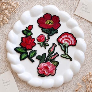 Puede incluir: Una colección de cinco parches bordados con motivos florales que representan varias rosas rojas y rosas con hojas y tallos verdes. Los parches están dispuestos sobre un plato blanco festoneado. Los diseños incluyen flores y capullos.