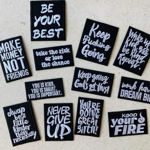 May include: A collection of black embroidered patches with white text. The patches feature motivational phrases like "BE YOUR BEST", "MAKE MONEY NOT FRIENDS", and "NEVER GIVE UP". These patches are ideal for customizing clothing or accessories.