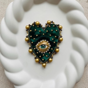 May include: A heart-shaped brooch with a dark green velvet base, adorned with gold beads and a central eye design. The eye features a green iris, surrounded by a gold frame and clear crystals. The brooch is set against a white, textured background.