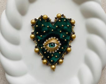 Op de afbeelding: Een hartvormige broche met een donkergroene fluwelen basis, versierd met gouden kralen en een centraal oogontwerp. Het oog heeft een groene iris, omgeven door een gouden frame en heldere kristallen. De broche staat op een witte, getextureerde achtergrond.