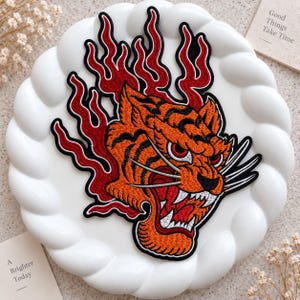 Pode incluir: Um emblema bordado apresentando uma cabeça de tigre laranja com listras pretas, olhos vermelhos e presas brancas, cercado por chamas vermelhas estilizadas com contornos pretos. O design é detalhado com bigodes brancos e uma borda preta.