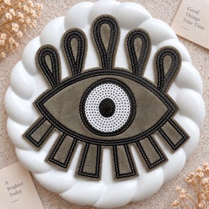 May include: An olive green felt patch shaped like an eye with black stitched outlines and a central white sequined iris with a black pupil. The design features five upward-pointing loops and five downward-pointing rectangular lashes in olive green felt.