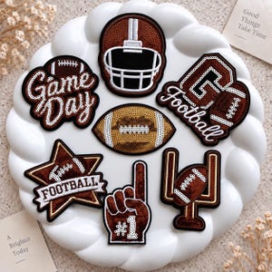 May include: A collection of seven football-themed patches featuring sequins in brown, white, and gold. Designs include a helmet, a football, a star with the word Football, a number one foam finger, goal posts, and text reading Game Day and Football.